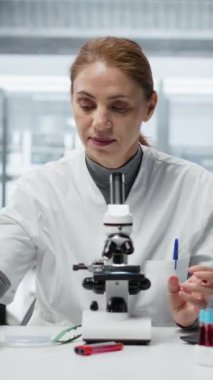 Laboratuvardaki dikey video tarih teknisyeni, yapısal bozukluklar ya da safsızlıklar için ince cam kaydırağını inceliyor. Laboratuvarda çalışan bir kadın mikroskop kullanarak görsel kalite kontrolü yapıyor.