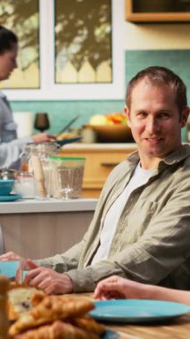 Dikey Videolu Neşeli Anne, çocuğuna ve ergen kızına masada yemekle servis yapıyor. Pastırma ve yumurtaları tabağa koyuyor. Aile, sabah ışığında kahvaltı için mutfak masasının etrafında toplandı.