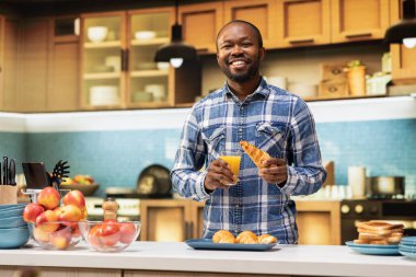 Afro-Amerikan bir adamın portresi. Elinde bir bardak portakal suyu ve mutfakta gülümseyen bir kruvasan tutuyor. Sabah rutini rahat ev atmosferinde sakinliği ve mutluluğu ifade eder..