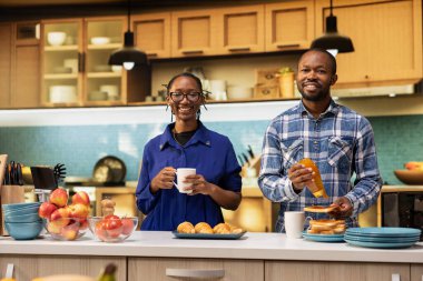 Afro-Amerikan aşıkların portresi mutfağa yakın duruyor, sabah kahvesinin ve ballı el yapımı pastanın tadını çıkarıyor. Lezzetli bir ev kahvaltısı sunmadan önce gülümseyen neşeli çift.