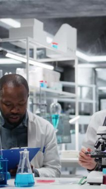 Meslektaşım panoya teşhis bulgularını yazarken dikey video patolojisti biyopsi slaytını inceliyor. Tıbbi uzmanlar klinik laboratuarında petri kabı hastalıklarını belirlemek için birlikte çalışıyorlar.