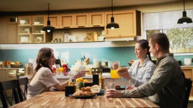 Aile zengin bir kahvaltı için mutfak masasının etrafında toplandı, hamur işi ve taze portakal suyunun tadını çıkardılar. Ebeveynler ve kızları arasındaki yemek sırasında neşeli sabah röfleleri. Kamera A.