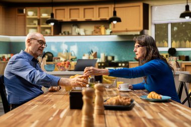 Olgun bir çift kahvaltı servisi yapıyorlar. Hamur işi, sıcak kahve, parlak mutfak masası. Sakin bir sabah rutini ve rahat bir emeklilik hayatı içinde sohbet ve kaynaşma..