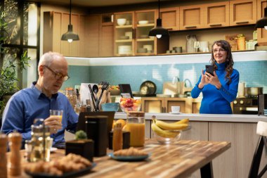 Apartman mutfağında akıllı telefondan kayan ve rahat ev işleri yapan yaşlı doğal kadın portresi. Yaşlı yetişkin neşeli yaşam tarzını yansıtıyor ve emekli hayatı tatmin ediyor..