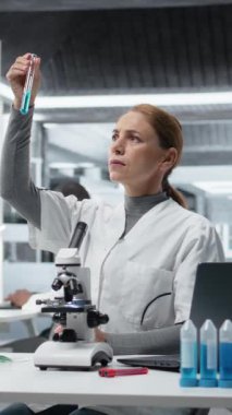 Dikey video laboratuvar teknisyeni test tüpündeki mavi sıvı kimyasal örnekleri inceliyor. Araştırma tesisi çalışanı yeni bileşik içeren şişeyi tutuyor. Tıbbi araştırma ve araştırma yapıyor. Kamera A.