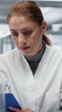 Dikey video laboratuarı çalışanı mikroskop ile biyopsi slaytını inceliyor, panoda teşhis bulgularını belgeliyor. Orta yaşlı bir kadın klinik laboratuarında hastalık belirtilerini tespit edip not alıyor.