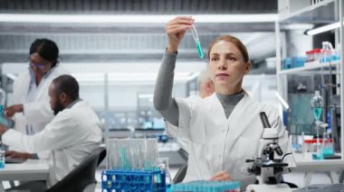 Kalite kontrol analisti test tüpündeki mavi sıvı berraklığı inceliyor ve laptopa veri giriyor. Endüstriyel ürün üretim süreci için kimyasal saflık standartlarını onaylayan laboratuvar teknisyeni, kamera B