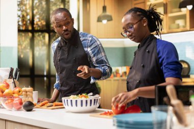 Kesme tahtasında kırmızı dolma biber doğrayan Afro-Amerikalı kadın erkek arkadaşıyla mutfakta taze bir salata hazırlıyor. Kız öğle yemeği için organik sebze ve malzeme hazırlıyor..
