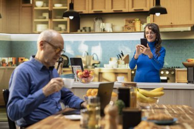 Yetişkinlerin akıllı telefonlarıyla internete bakması, mutfakta gülümsemesi, kahveyle rahatlaması ve huzurlu emeklilik hayatının tadını çıkarması. Emeklilik, pozitif yaşam tarzı ve doğal ev hayatı..