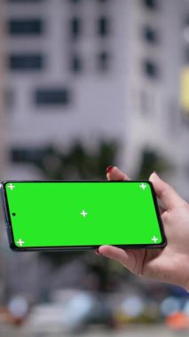 Dikey video krom anahtar akıllı telefon kulaklıklı ve sırt çantalı genç bir yetişkin tarafından tutuluyor. Hippi kız kalabalık iş yeri sokaklarında yürüyor. Farklı yaşlarda sıradan yayalar.