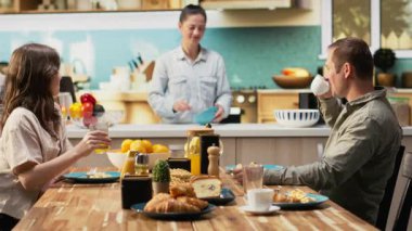 Anne sabah ışığında kahvaltı hazırlarken aile masada kruvasan, meyve ve portakal suyuyla bekliyor. Ebeveynler ve küçük kızları birlikte gülüyorlar, tatlım. Kamera B.