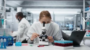 Tıbbi laboratuvarda cerrahi maske takan bir biyolog hücresel analiz için bilimsel mikroskop kullanıyor. Laboratuvarda yüksek teknoloji optik cihaz teçhizatıyla test edilen kadın bilim adamı, kamera B