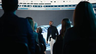 Sahnede endüstriyel makineler için türbin sunan siyahi konuşmacı, uluslararası teknoloji konferansı sırasında mekanik ekipman görselleri. Konferans salonunda panel tartışması.