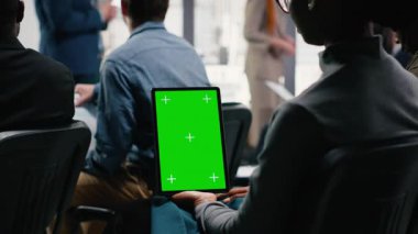 Dik yeşil ekran tableti kurumsal iş forumunda, sahnede konuşma yapan kişi tarafından tutuldu. Beyaz yakalı işçilerden oluşan çeşitli seyirciler panel etkinliği sırasında dinliyorlar. Kamera B.