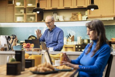 Yaşlı yetişkin, dizüstü bilgisayarında ailesiyle tatlı bir bağ kurmak için uzaktan video kullanıyor. Yaşlı adam mutfakta oturuyor ve internette telekonferansa katılıyor, sakin bir emeklilik yaşam tarzını yansıtıyor..