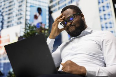 Afro-amerikan genç yetişkin üzgün ve bitap bir halde bir bankta oturuyor, hoşnutsuz ve endişeli bir şekilde dizüstü bilgisayarında oturuyor. Şehir mimarisi ve şehir yaşamının ritmiyle çevrili..