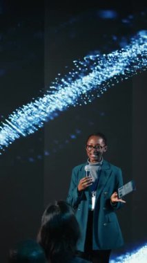 Dikey Video Siyah kadın konuşmacı mikrofon kullanarak uluslararası konferans için profesyonel iş sunumu yapıyor. Girişimcilik konferansı sırasında konferans salonundaki katılımcılar