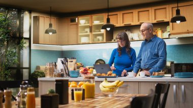 Olgun evli çiftler dizüstü bilgisayarlarında geziniyorlar ve sakin bir şekilde gülümsüyorlar, pozitif bir emeklilik hayatı ve doğal bir sabah rutini sergiliyorlar. Memnuniyet verici bir yaşam tarzı. Kamera A.