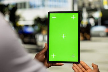 Şehir parkında bir bankta oturan siyahi adamın tablet üzerinde güvenle online görevleri yazıp yönettiği dikey izole ekranı kapatın. Mimari ve canlı şehir hayatı.