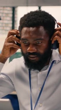 Dikey Video Siyah çalışanı, modern ofiste müzik eşliğinde kulaklık takarak, çalışma alanında neşe ve neşe içinde eğleniyor. Stres atmak için şarkı dinleyen bir adam. Dengeli kurumsal yaşam tarzı.