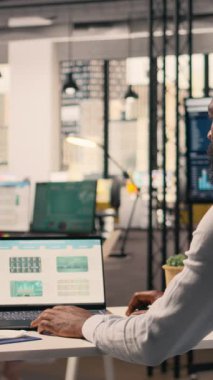 Dikey Video Siyah erkek profesyonel tahmin raporlarını ve görsel verileri analiz ediyor. Masasında dizüstü bilgisayarı olan kurumsal ofis ortamını. Analiz paneli planlama ve görev için dizüstü bilgisayarda görüntülendi