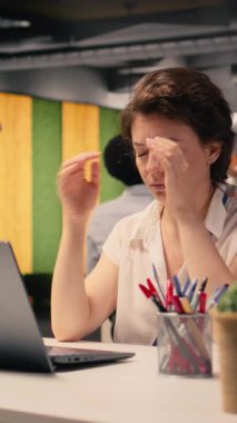 Baş ağrısı ve göz yorgunluğu yaşayan, kurumsal ofiste çalışan, gözle görülür yorgunluk ve stresle şakakları ovuşturan dikey video iş kadını. Migren ve iş geriliminden dolayı bulanık görme