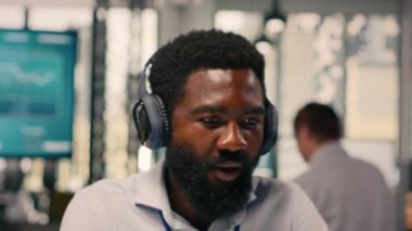 Neşeli siyahi adam kablosuz kulaklıklardan müzik listesi dinliyor, çizelgeleri ve grafikleri dizüstü bilgisayardan inceliyor. Rahatlamış işçi eğlenceli müzikle çoklu görev, modern ofiste stres atımı.