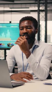 KPI ölçümlerini ve modern ofis çalışma alanındaki sezgilerini analiz eden Afrikalı Amerikalı iş adamının dikey video portresi. Görsel veriler, tahmin raporları, stratejik planlama ve küresel planlama için dizüstü bilgisayar kullanımı