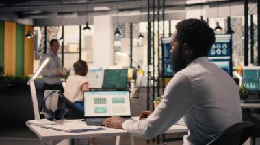 Siyahi erkek profesyonel tahmin raporlarını ve görsel verileri analiz ediyor. Ofis ortamında dizüstü bilgisayarı var. Analitik gösterge paneli planlama ve görev yönetimi için dizüstü bilgisayarda görüntülendi.