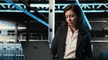 Veri merkezi yazılım geliştiricisi dizüstü bilgisayarla sunucu yükü dengelemeyi optimize ediyor. Verimliliği artırmak için sistem parametrelerini masadaki sunucu çiftliğindeki kadın, kamera B