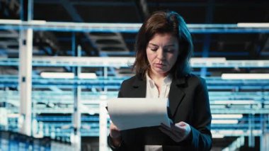 Veri merkezi yönetimi evrakları okuyor, mavi kablo ağ donanım donanımlarını inceliyor. Sunucu çiftliğinde çalışan kadın AR altyapı görselleştirmesi ile çevrili belgeleri analiz ediyor, kamera B