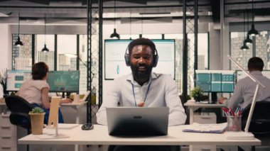Modern ofiste kulaklıklarla video konferansına katılan siyahi bir profesyonel. Çevrimiçi bilgilendirme, şeffaflık ve iletişim sırasında çizelgeleri ve veri analizlerini gösteren iş yeri.