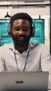 Dikey Video Neşeli Zenci adam kablosuz kulaklıklarda müzik listesi dinliyor, grafikleri ve grafikleri dizüstü bilgisayardan inceliyor. Rahatlamış işçi eğlenceli müzikle çoklu görev, modern ofiste stres atımı.