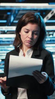 Dikey Veri Merkezi Yöneticisi altyapı durumunu pano dosyaları ve holografik görsellerle onaylıyor. Denetim çizelgeleri ve mavi kablo simülasyonu kullanarak sunucu odası çalışanı