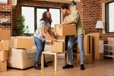 Gülümseyen sevgili, yeni alınmış bir dairede karton kutular taşıyor ve düzenliyor. Ev taşınması ve yeni bir hayata başlama süresince takım çalışması ve beraberlik gösteren çift.