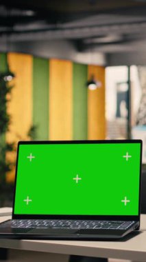 Boş şirket masasındaki dizüstü bilgisayarda dikey video yeşil ekran modeli modern ofis alanındaki krom anahtar görüntüsü. Modern mobilyalar, ofis malzemeleri ve insan olmayan parlak bir iş ortamı..