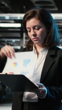 Dikey video okuma evrakları, mavi kablo örgüsü ve veri merkezindeki donanım donanımlarını denetleyen yönetici. Belgeler analiz ediliyor, AR altyapı görselleştirme ve sunucu çiftliğinde çalışan kadın, kamera B