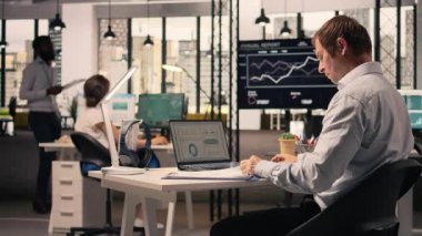 Erkek ofis çalışanı masadaki görevleri bağımsız olarak çözüyor, görsel verileri, grafikleri ve grafikleri gözden geçiriyor. Üretkenlik ve küresel iş planlaması için tahmin raporları inceleniyor.