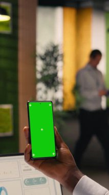 Ofiste yalnız çalışan Afrikalı bir Amerikalı tarafından tutulan dikey video krom anahtar telefon, çalışma alanında raporlar ve KPI ölçümleri tahmin ediyor. Grafikler, grafikler ve içgörüler organizasyonu destekler