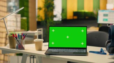 Boş şirket masasındaki dizüstü bilgisayarın yeşil ekran modeli modern ofis alanındaki krom anahtar görüntüsü. Modern mobilyalar, ofis malzemeleri ve insan olmayan parlak bir iş ortamı..
