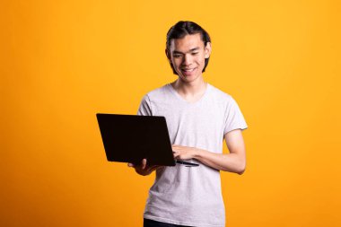 Asyalı adam mutlu bir şekilde dijital cihaza sahip, e-postalara cevap veriyor ve interneti tarıyor. İletişim ve çevrimiçi işbirliği için dizüstü bilgisayar kullanan neşeli serbest yazar, neşeli bir ifadeyle ayakta duruyor.