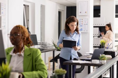 Kararlı iş kadını masanın başında duruyor, panodaki verileri dizüstü bilgisayardaki dijital raporlarla karşılaştırıyor. Analizleri, proje güncellemelerini ve şirket performans detaylarını inceleyen şirket danışmanı.