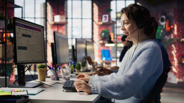 Ofisteki mutlu çağrı merkezi ajanı soruları cevaplayarak, sorunları çözerek müşterilere yardım ediyor. Müşteri hizmetlerindeki gülümseyen kadın bilgisayarın önünde müşterilerle konuşmak için kulaklık kullanıyor, kamera B