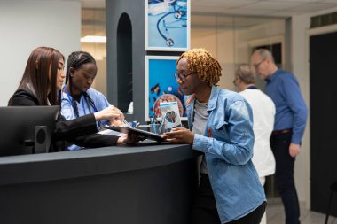 Asyalı yönetici hastane lobisindeki tıbbi belgeleri tamamlamak için siyah kadına yardım ediyor. Profesyonel resepsiyonist ve kadın hasta klinik resepsiyonunda sohbet ediyorlar..