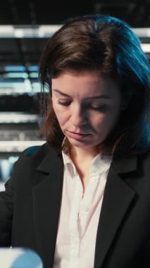 Altyapı durumunu doğrulayan dikey video, pano dosyaları ve holografik görsellerle veri merkezinde yönetici. Denetleme ekipmanı, kablo simülasyonu ve denetim kullanarak sunucu odasında çalışan