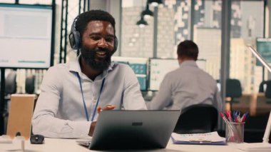 Modern ofiste kulaklıklarla video konferansına katılan siyahi bir profesyonel. Çevrimiçi bilgilendirme, şeffaflık ve iletişim sırasında çizelgeleri ve veri analizlerini gösteren iş yeri.