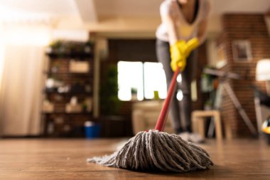 Sarı lastik eldivenli bir kadın oturma odasının zeminini paspaslıyor ve ahşap zemine odaklanıyor. Kadın ev sahibi ev işlerini yapıyor, temizliği sağlıyor ve bakımını yapıyor.