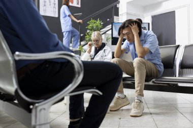 Endişeli Asyalı adam ellerini başının üstüne koyup hastane bekleme odasında teşhis sonuçlarını bekliyor. Duruşu, gergin sağlık durumundaki duygusal stres ve belirsizliği yansıtıyor..