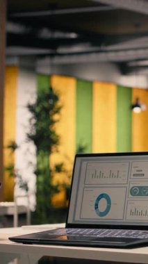 Laptop ekranında çalışan dikey video performans ölçümleri boş açık ofiste malzeme ve aletlerle, modern iç dekorasyonda mobilya ve doğal ışıkla dekore edilmiş masa düzeneği. Çalışma ortamı.