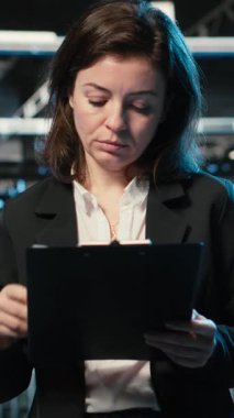 Altyapı durumunu doğrulayan dikey video, pano dosyaları ve holografik görsellerle veri merkezinde yönetici. Denetleme ekipmanı, kablo simülasyonu ve denetim kullanarak sunucu odasında çalışan